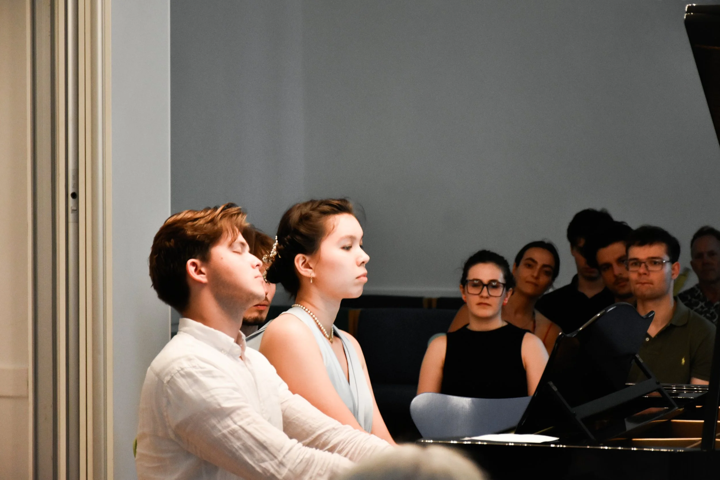 Mehrere Menschen sitzen in einem Raum, einige mit geschlossenen Augen, während Hannah Trapp und Benjamin Britten am Klavier sitzten und konzentrativ spielen.