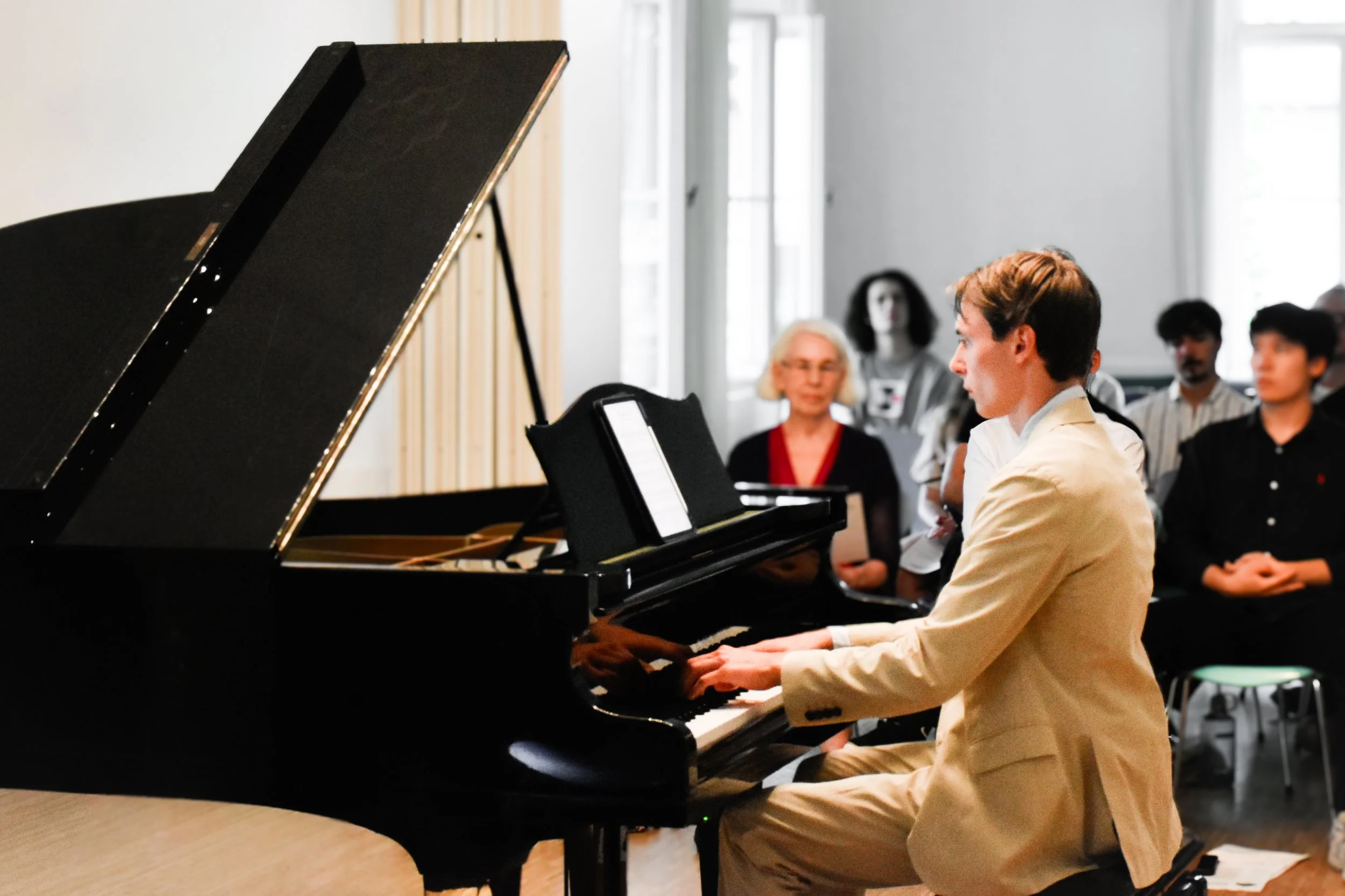 Ein junger Mann spielt Klavier in einem Raum, während mehrere Menschen im Hintergrund zuhören.