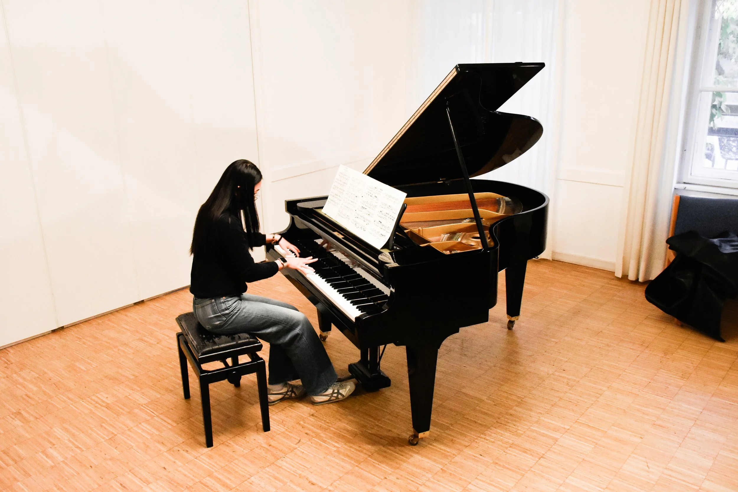 Eine junge Frau spielt Klavier in einem Musikraum, sitzt auf einem Hocker, während das Klavier offen ist, um die Saiten zu zeigen.
