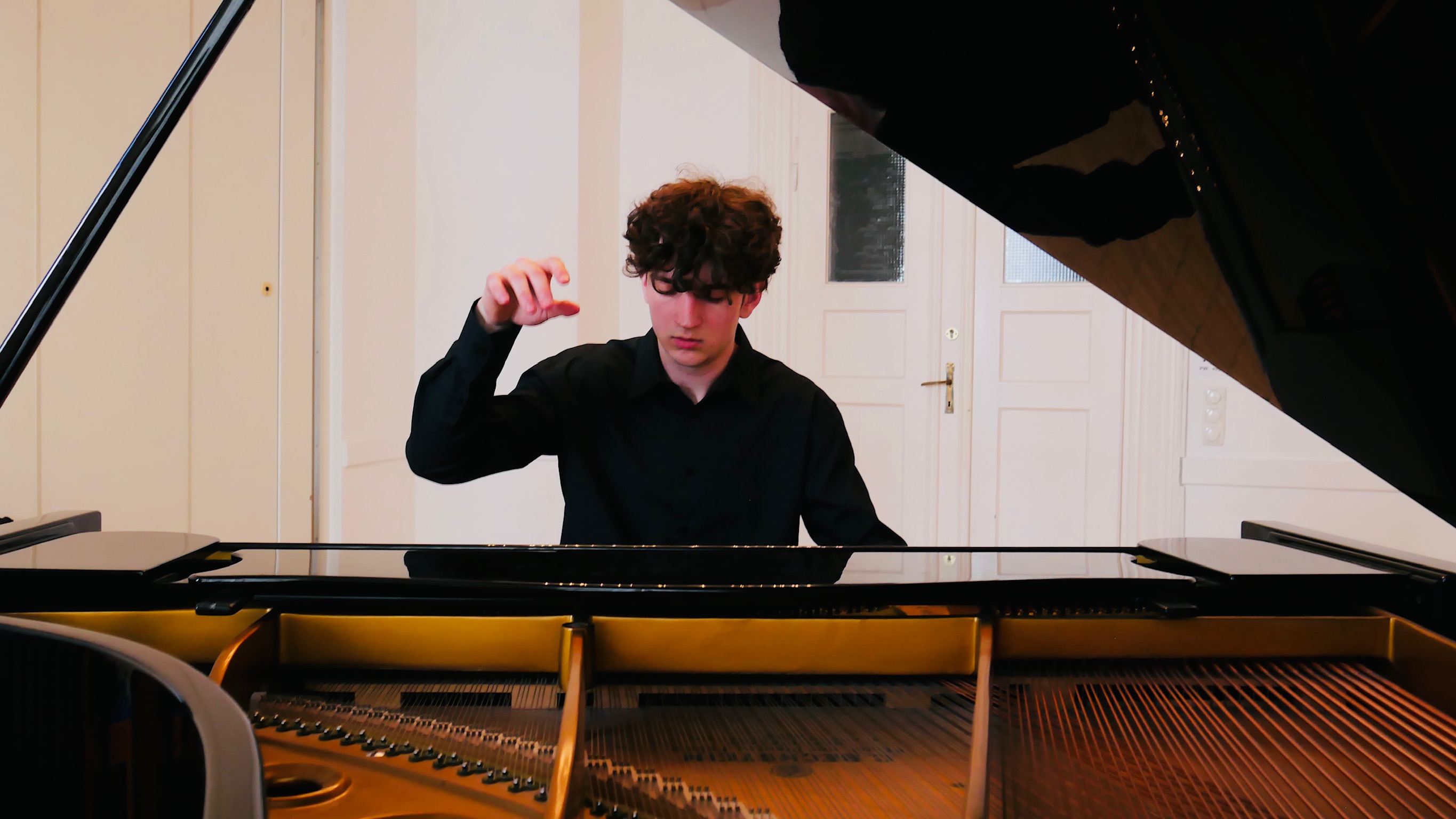 Ein junger Mann mit dunklen Locken spielt einen Flügel, die rechte Hand über der Klaviatur erhoben.